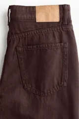 Pantalón tipo jean de corte wide leg y tiro medio, confeccionado en denim de algodón rígido color marrón oscuro. Presenta diseño clásico de cinco bolsillos, cierre frontal con botón y cremallera, y piernas amplias con caída recta.