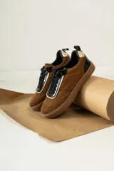 Championes urbanos de gamuza color marrón, con detalles en charol negro y bordes dorados. Presentan cordones negros y suela de goma al tono.