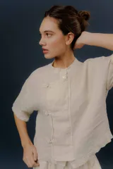 Blusa de lino color crudo, con cuello mao, mangas cortas y cierre frontal cruzado con nudos de tela y botones.