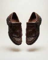 Mocasín de cuero color bordó con textura trenzada en la puntera. Presenta una solapa desmontable con flecos y detalle de perforaciones, junto con una hebilla lateral metálica. Diseño versátil que permite transformar el calzado.