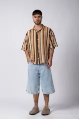 Camisa de manga corta con cuello solapa y cierre frontal de botones. Presenta un diseño de rayas verticales en tonos marrones, beige y negro, con una textura tejida y corte holgado.