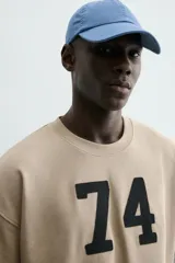 Sudadera de manga corta color beige, corte relajado (relaxed fit) con cuello redondo y número "74" bordado en negro en el frente.