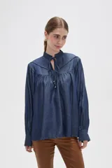 Blusa de lyocell en tono azul denim, con diseño holgado y frunces en el canesú. Presenta cuello alto con volados, cierre frontal mediante cordón ajustable y mangas largas con puños abotonados.