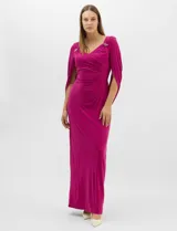 Vestido largo de seda fría color fucsia, con escote cruzado drapeado y apliques de pedrería en los hombros. Presenta mangas largas abiertas estilo capa y un drapeado lateral en la cintura que estiliza la silueta.