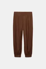 Pantalón de tiro alto en color beige, con diseño de pierna holgada y puños elásticos con cierre de botón en el bajo. Presenta cierre frontal con cremallera y doble botón en la cintura, además de bolsillos laterales.