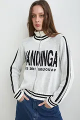 Campera tejida de algodón en color blanco con detalles en negro, de cuello alto con cierre frontal y franjas decorativas en las mangas. Cuenta con diseño estampado en el frente.
