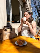 Cartera tipo tote de cuero color beige, con doble asa corta marrón y cierre superior con cremallera. Tiene un bolsillo frontal con cremallera y forro interior de tela con un bolsillo.