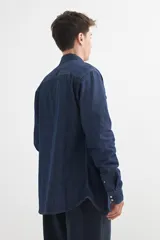 Camisa de denim azul con cuello clásico, cierre frontal con botones a presión, dos bolsillos con solapa abotonada en el pecho y mangas largas con puños abotonados.