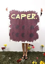 Pañuelo cuadrado de tela con estampado floral en tonos bordó sobre fondo crudo y la palabra 'CAPER.' en color amarillo en la parte superior.