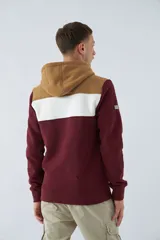 Buzo canguro de felpa color borgoña, con capucha ajustable con cordones color beige, bolsillo tipo canguro y logo estampado en el pecho. Tiene una franja horizontal blanca en el pecho y puños y cintura acanalados.