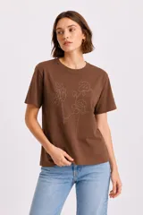 Remera de algodón color marrón, de corte holgado y cuello bote, con diseño floral realizado mediante aplicación de studs metálicos en el frente.