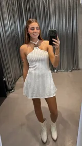 Vestido corto de color blanco con textura acanalada y diseño de cuello halter con detalle metálico circular en el centro. Presenta un corte ajustado al cuerpo y una caída fluida hacia el ruedo.
