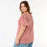 Camiseta rosada de manga corta con escote en V, confeccionada en tejido texturizado con detalles de encaje y macramé en los hombros.