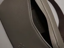 Riñonera de poliuretano color gris topo, con textura granulada y logo 'CK' grabado en el frente. Posee una correa ajustable de tela marrón oscuro.