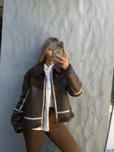 Campera tipo gamulán de cuero sintético en color marrón oscuro, con interior y detalles de cuello y puños en corderito sintético al tono. Presenta cierre frontal, correas ajustables en el cuello y diseño con costuras reforzadas.