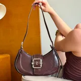 Cartera tipo baguette de color marrón con acabado texturizado. Presenta una hebilla decorativa central con la letra Z en metal plateado, cierre principal con cremallera y correa de hombro ajustable.