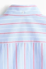 Camisa de corte relajado confeccionada en algodón, con diseño de rayas verticales en tonos azul, blanco y rojo. Presenta cuello americano, cierre frontal con botones, bolsillo en el pecho y mangas largas con puños abotonados.