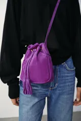 Mini bandolera de piel color morado, de diseño tipo saco con cierre fruncido y cordón ajustable con borlas en el extremo. Cuenta con asa larga ajustable y detalles metálicos dorados.