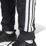 Pantalón de jogging Adidas modelo M 3s Ft Tc Pt, color negro con las tres tiras características en los laterales de color blanco y el logo de la marca bordado en la pierna izquierda. Cuenta con cintura elástica con cordón de ajuste y puños en los tobillos.