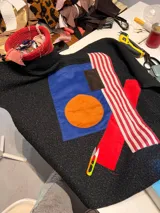 Campera de paño negro con diseño de collage artístico en el frente, compuesto por parches de tela en colores azul, naranja, rojo y blanco con rayas. Presenta un corte holgado y diseño artesanal único.
