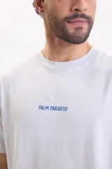 Remera blanca de manga corta con gran estampado azul en la espalda que representa un paisaje tropical con palmeras, mar y aves.