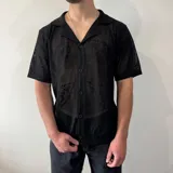 Camisa de tejido de punto calado color negro, con cuello solapa, cierre frontal de botones y mangas cortas.