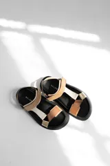 Sandalias planas con suela negra, tiras anchas de cuero color beige y blanco, y detalle de apliques dorados.