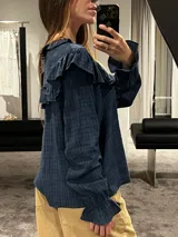 Camisa blanca de textura rugosa con volados en el pecho y hombros, cuello redondo con lazos de ajuste, cierre frontal con botones y mangas largas con puños con volados.