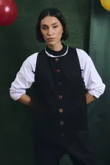 Mono largo de color negro con cierre frontal de botones vintage de distintos colores. Presenta un diseño sin mangas, con bolsillos laterales y una cinta para herramientas en la parte trasera.