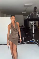 Vestido midi de color marrón, con diseño de cuello halter y escote drapeado. Presenta un corte ajustado con frunces laterales y una abertura pronunciada en la pierna que le otorga un estilo asimétrico.
