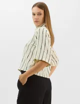 Camisa de corte holgado y silueta recta, con estampado de rayas verticales irregulares en color negro sobre fondo crema. Presenta cuello camisero clásico, mangas cortas amplias y cierre frontal con botones.