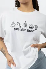 Remera blanca de algodón con estampa frontal en color negro que incluye iconos de café y la frase 'More coffee, please!'. Presenta cuello redondo y mangas cortas.