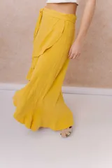 Falda larga envolvente de color mostaza, con diseño asimétrico, lazo ajustable en la cintura y volados en cascada.