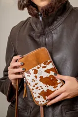 Pocket bag de cuero vacuno con diseño combinado en gamuza color tostado y cuero con pelo estampado animal print tipo vaca. Cuenta con bolsillo frontal con detalle de bordado metálico, cierre superior y correa desmontable.