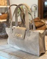 Cartera tipo tote bag de gamuza en color beige, con diseño rectangular, doble asa de hombro y cierre frontal con broche metálico dorado.