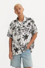 Camisa de manga corta con cuello tipo cubano, confeccionada en tela estampada con diseño tropical de palmeras y flores en blanco y negro. Presenta cierre frontal con botones y un bolsillo en el pecho.