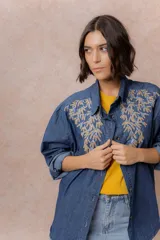 Camisa de denim azul con bordados florales en tonos amarillos y celestes sobre los hombros y el pecho. Presenta cuello clásico, cierre frontal con botones y mangas largas con puños.