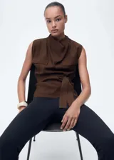 Blusa marrón sin mangas con cuello drapeado, trabillas, dos bolsillos de solapa en el pecho y cinturón desmontable. Cierre de botón en la parte posterior y diseño recto.