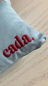 Almohada pequeña de tela celeste con la palabra "cada." bordada en rojo.