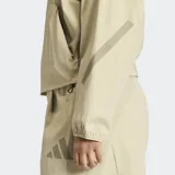 Campera tipo bomber Adidas Z.N.E. de corte holgado, color beige, con cierre frontal completo, cuello tipo béisbol y logo de las 3 barras en caucho mate en el pecho. Confeccionada en tejido ripstop con protección UV, presenta puños elásticos y diseño minimalista.