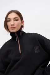 Buzo oversize de felpa con friza, color negro, con cuello alto y cierre frontal parcial metálico. Presenta bolsillos laterales, tajos en los costados y un diseño con espalda más larga. Incluye un pequeño logo bordado en el pecho.