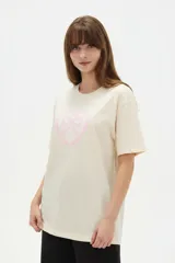 Remera de algodón color beige con estampado frontal de un corazón rosado que contiene un moño. Tiene cuello redondo y mangas cortas.