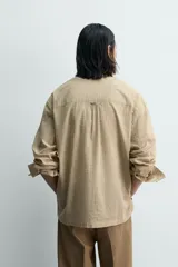 Camisa de hombre de manga larga, color beige con micro-estampado a cuadros en tonos marrones. Presenta cuello solapa abotonado (button-down), un bolsillo de parche en el pecho y cierre frontal de botonadura.