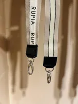 Correa para cartera de cinta tejida en tonos beige y crema, con detalles de costuras negras y logo impreso. Incluye terminales de cuero sintético negro y ganchos metálicos tipo mosquetón plateados para sujeción.