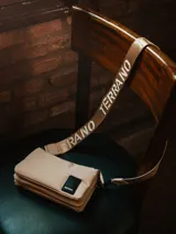 Bandolera de diseño rectangular y estilo acolchado, color beige, con correa larga ajustable que presenta el nombre de la marca estampado repetidamente. Incluye un parche rectangular negro con el logo de la marca en el frente.