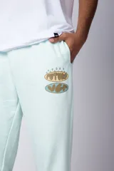 Pantalón jogger de corte oversize confeccionado en tela rústica color verde agua claro. Presenta una estampa gráfica localizada en el muslo izquierdo con diseño de óvalos y estrellas.