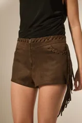 Shorts de gamuza color marrón oscuro, de tiro medio y corte al cuerpo. Presenta detalles de cordones trenzados en los bordes superiores y en los bolsillos delanteros, además de flecos largos cayendo desde el lateral derecho.