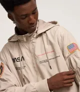Campera rompevientos color beige con capucha, cierre frontal y bolsillos laterales. Presenta estampado de la NASA en el pecho, parche circular en la manga derecha y bandera de Estados Unidos en la manga izquierda.