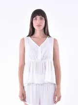 Blusa sin mangas de color blanco, con escote en V y cierre frontal parcial con botones. Presenta un corte tipo peplum con fruncido en la cintura que genera un ligero vuelo.