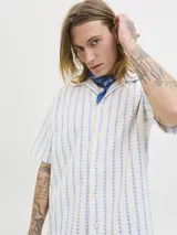 Camisa de manga corta estilo resort, confeccionada en tejido jersey de punto con diseño de rayas verticales en tonos crudo y azul. Presenta cuello camisero y cierre frontal con botones.